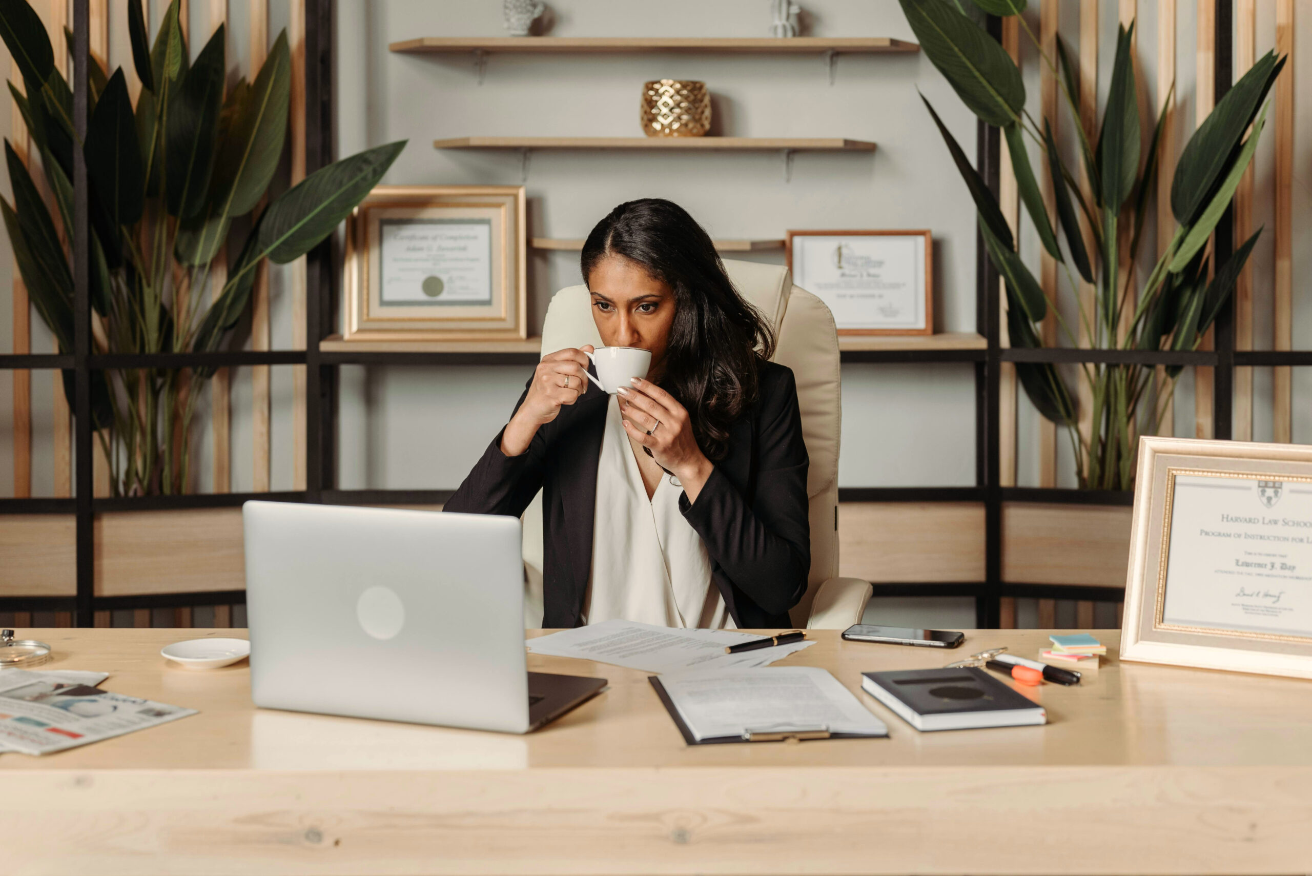 A well dressed woman with black hair sips coffee at her desk